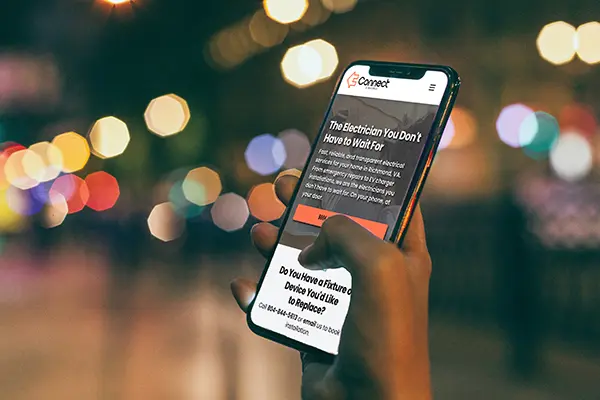 A hand holding a smartphone displaying the mobile homepage of Connect Electric, an electrical services website, against a blurry night city lights background.