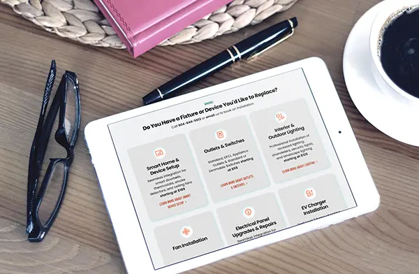 A white tablet on a wooden desk displaying the services page of a responsive electrician website, Connect Electric, alongside glasses, a pen, and a coffee cup.