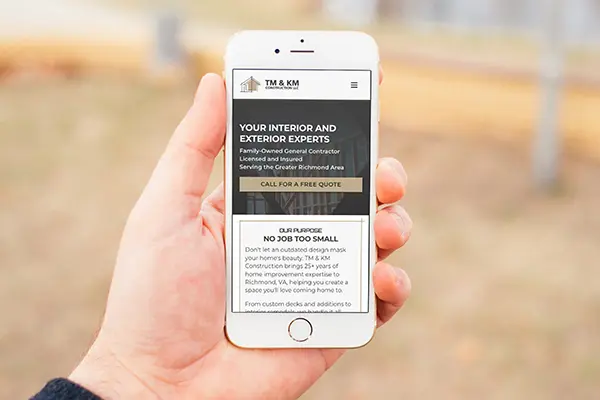 A person holding a white smartphone displaying the mobile-responsive version of the TM & KM Construction website. The screen shows the company logo and the headline 